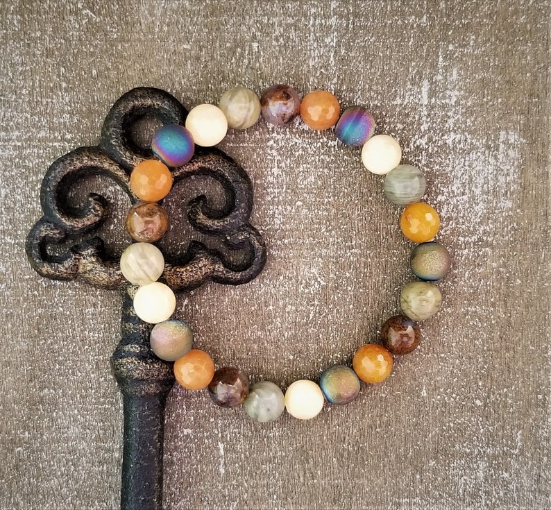 May include: A beaded bracelet with a variety of colors and textures, including brown, white, orange, and iridescent blue. The bracelet is lying on a wooden surface.