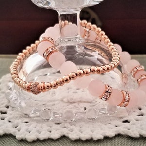 May include: Two bracelets, one with rose gold beads and the other with pink beads and clear crystals. The bracelets are stacked on top of each other.