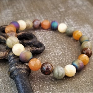 May include: A multi-colored beaded bracelet with a mix of brown, orange, white, and iridescent beads. The bracelet is lying on a wooden surface next to an antique key.