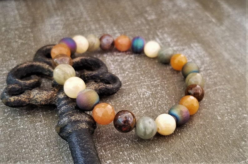 May include: A multi-colored beaded bracelet with a mix of brown, orange, white, and iridescent beads. The bracelet is lying on a wooden surface next to an antique key.