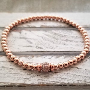 May include: A rose gold beaded bracelet with a single larger rose gold bead accented with small clear stones.