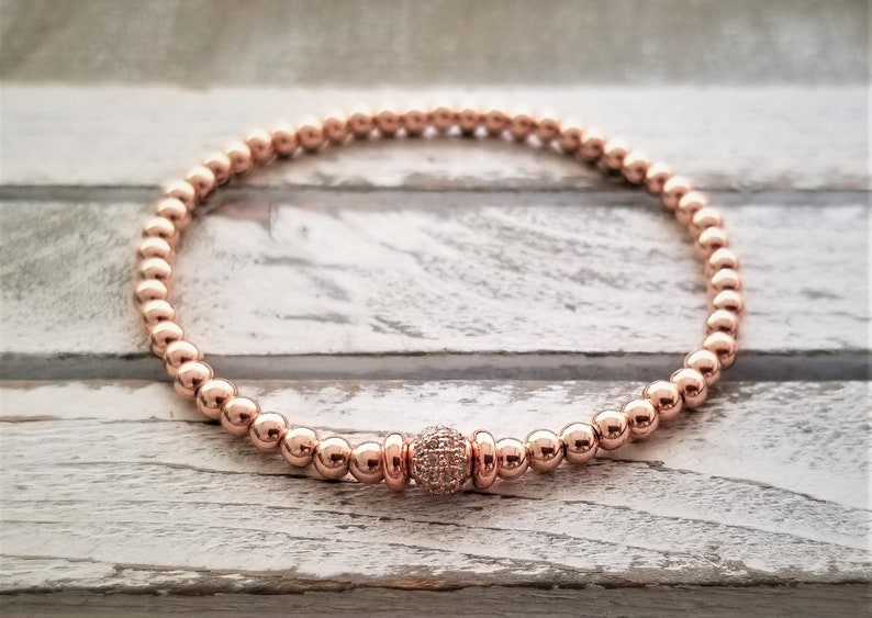 May include: A rose gold beaded bracelet with a single larger rose gold bead accented with small clear stones.