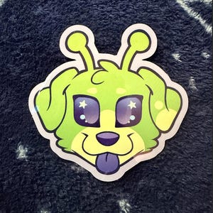 May include: A die-cut sticker featuring a cartoon alien-dog hybrid. The sticker has a lime green head with large, purple eyes containing white stars. The dog's tongue is a contrasting purple, and the sticker has a white border.