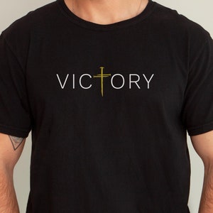 May include: Black t-shirt with a white graphic that says "VICTORY" with a gold cross in the middle of the word.