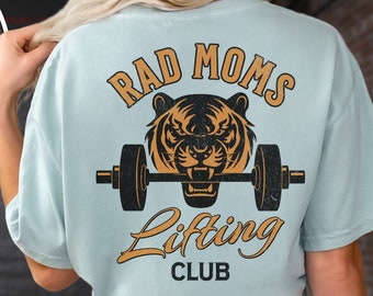 Może przedstawiać: Jasnoniebieska koszulka z grafiką przedstawiającą tygrysa podnoszącego ciężary. Tekst na plecach brzmi "RAD MOMS Lifting CLUB" w kolorze pomarańczowym i czarnym. Przód koszulki ma mniejszą wersję grafiki tygrysa i ciężarów.