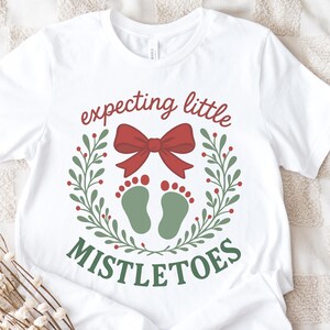 May include: White t-shirt with a festive design. The shirt features the text "expecting little mistletoes" in red and green, with a red bow and green baby footprints. A holiday-themed tee for expecting parents.