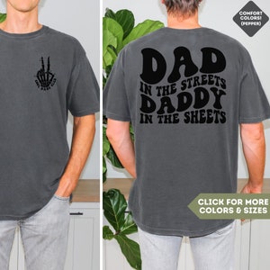 May include: A dark gray t-shirt with a retro-style graphic on the back that says "Dad in the streets Daddy in the sheets". The front of the shirt has a small graphic of a hand making a peace sign with the words "Cool Dad's Club".