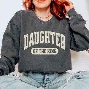 May include: A dark gray sweatshirt with the text "Daughter of the King" in a white arch design.