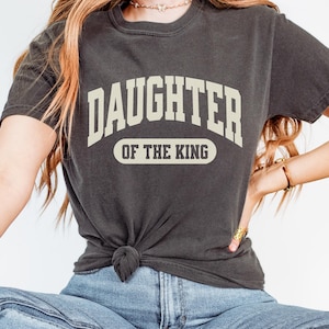 May include: A dark gray t-shirt with the text "Daughter of the King" in a white arch design.