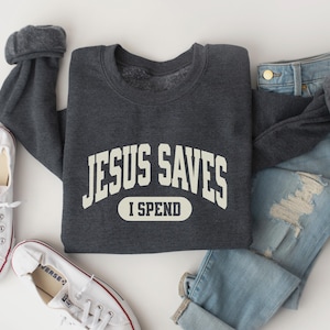 May include: A dark gray sweatshirt with the text "JESUS SAVES I SPEND" in a white arch design. The sweatshirt is laid out on a white surface with a pair of white sneakers and a pair of light blue jeans.