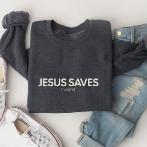May include: A dark gray sweatshirt with the text "JESUS SAVES I Spend" in white lettering.