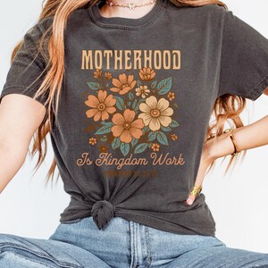 May include: A dark gray t-shirt with a floral design and the text "Motherhood is Kingdom Work" printed on the front.