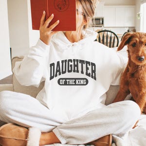 May include: A white hoodie with the text "Daughter of the King" in black lettering. The hoodie is worn by a person sitting on a couch with a dog.