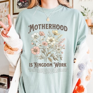 May include: Light blue t-shirt with a floral design and the text "MOTHERHOOD IS KINGDOM WORK". The text is in brown and the flowers are in shades of pink and white. The quote below the text reads "SHE IS CLOTHED WITH STRENGTH AND DIGNITY. SHE CAN LAUGH AT THE DAYS TO COME. SHE SPEAKS WITH WISDOM, AND FAITHFUL INSTRUCTION IS ON HER TONGUE. SHE WATCHES OVER THE AFFAIRS OF HER HOUSEHOLD AND DOES NOT EAT THE BREAD OF IDLENESS. - PROVERBS 31:25"