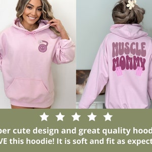 May include: Pink hoodie with a kettlebell graphic on the front and "MUSCLE MOMMY" text on the back. The hoodie has a drawstring hood and a kangaroo pocket.