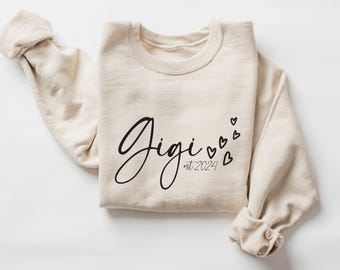 May include: A beige sweatshirt with the text "Gigi est. 2024" in black script font. The text is surrounded by small black hearts.