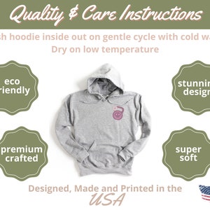 May include: A gray hoodie with a pink design on the front. The hoodie is made in the USA and is eco-friendly, premium crafted, super soft, and has a stunning design. The care instructions are to wash the hoodie inside out on a gentle cycle with cold water and dry on low temperature.