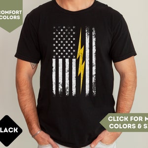 May include: Black t-shirt with a distressed American flag design featuring a yellow lightning bolt. The text "COMFORT COLORS" is visible in the top left corner of the image.