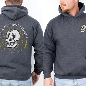 May include: A dark gray hooded sweatshirt with a skull graphic and the text "EVERYTHING HURTS AND I'M  HUNGRY" on the back. The front of the sweatshirt has a smaller skull graphic on the chest.