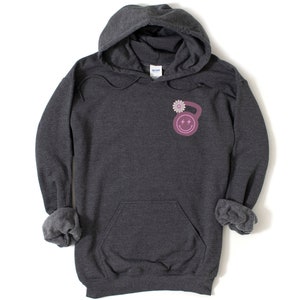 May include: A dark gray hooded sweatshirt with a pink and white kettlebell graphic on the front. The kettlebell has a smiley face with lightning bolts for eyes and a flower on top.