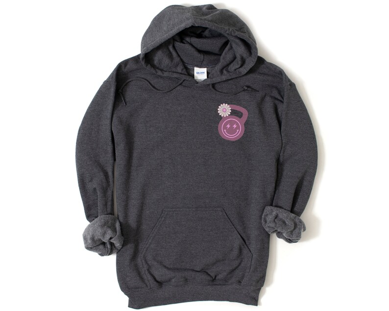May include: A dark gray hooded sweatshirt with a pink and white kettlebell graphic on the front. The kettlebell has a smiley face with lightning bolts for eyes and a flower on top.
