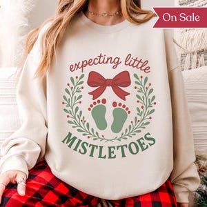May include: A sand-colored sweatshirt with the text "expecting little mistletoes" in red and green. The design includes a red bow, green footprints, and a wreath of green leaves. A red "On Sale" banner is visible.