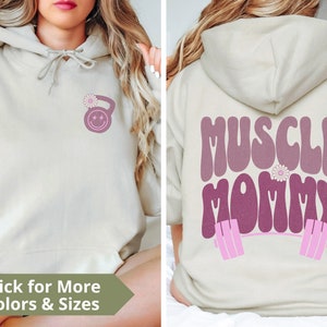 May include: A light beige hoodie with a pink and white flower and a pink dumbbell with a smiley face on the front. The back of the hoodie has the text "MUSCLE MOMMY" in pink with a pink barbell and a pink flower.