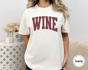 Peut inclure: Un t-shirt blanc avec le mot "WINE" imprimé en marron sur le devant. Le t-shirt est de coupe décontractée et a un col rond. Le mannequin porte le t-shirt en couleur ivoire.