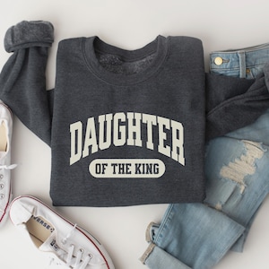 May include: A dark gray sweatshirt with the text "Daughter of the King" in a white arch design. The sweatshirt is laid out on a white surface with a pair of white sneakers and a pair of light blue jeans.