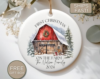 May include: White ceramic Christmas ornament with a red barn, snow-covered trees, and the text "FIRST CHRISTMAS ON THE FARM The Nelson Family 2024".