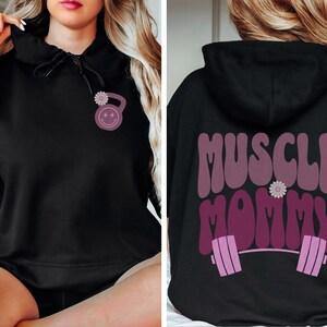 May include: Black hooded sweatshirt with a pink and white flower and a pink dumbbell with a smiley face on the front. The back of the sweatshirt has the text "MUSCLE MOMMY" in pink with a pink barbell and a pink flower.