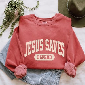 May include: Red sweatshirt with the text "JESUS SAVES I SPEND" in a vintage collegiate style font.