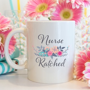 May include: White ceramic coffee mug with a floral design and the text "Nurse Ratched". The mug is surrounded by pink flowers.