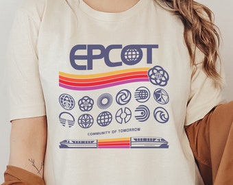 May include: A white t-shirt with a retro-style graphic of the Epcot logo and various park icons. The text "EPCOT" is in purple and the text "Community of Tomorrow" is in black. The graphic includes a rainbow stripe and a monorail.