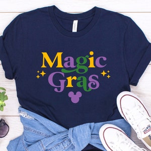 May include: A navy blue t-shirt with the text "Magic Gras" in yellow, green, and purple lettering. A purple Mickey Mouse head is below the text.