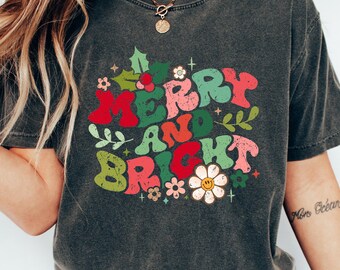 May include: A dark gray t-shirt with a retro-style graphic that says "Merry and Bright" in colorful letters surrounded by flowers.