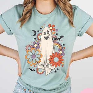 Peut inclure: Un t-shirt bleu clair avec un motif de fant&ocirc;me de style r&eacute;tro. Le fant&ocirc;me est blanc avec un visage souriant et est entour&eacute; de fleurs color&eacute;es, d'&eacute;toiles et de chauves-souris. Le design a une esth&eacute;tique vintage.
