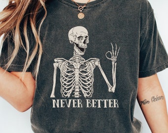 以下が含まれることがあります： 暗いグレーのTシャツに、骸骨のグラフィックと「Never Better」の文字がプリントされています。骸骨は笑顔で親指を立てています。