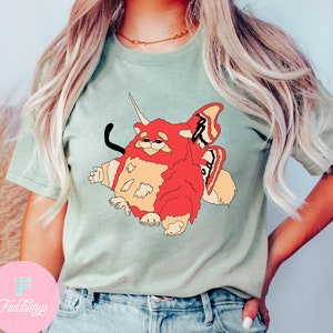 May include: A light green t-shirt with a cartoon illustration of a red, furry creature with butterfly wings. The creature has a horn on its head and a black tail.