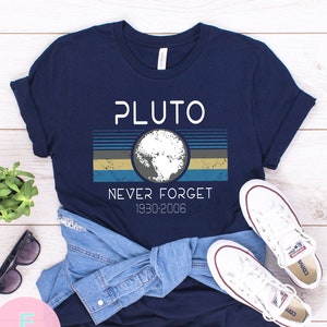 May include: A navy blue t-shirt with a white graphic of Pluto, a planet with a retro-style striped background. The text "Pluto Never Forget 1930-2006" is printed on the shirt.