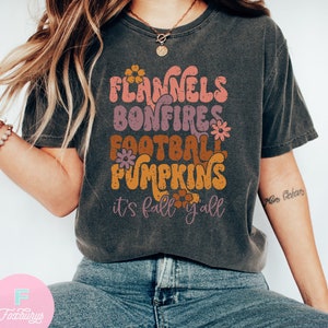 May include: A dark gray t-shirt with a retro-style graphic design featuring the words "Flannels, Bonfires, Football, Pumpkins, it's fall y'all". The graphic is in shades of orange, pink, and brown.