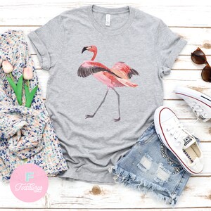 May include: A gray t-shirt with a pink flamingo illustration. The flamingo is standing on one leg with its wings spread out. The t-shirt is on a white wooden surface with a pair of denim shorts, white sneakers, and sunglasses.