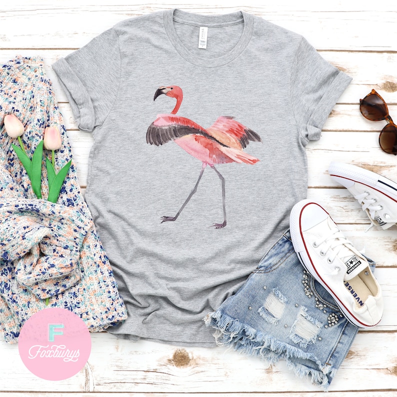 May include: A gray t-shirt with a pink flamingo illustration. The flamingo is standing on one leg with its wings spread out. The t-shirt is on a white wooden surface with a pair of denim shorts, white sneakers, and sunglasses.