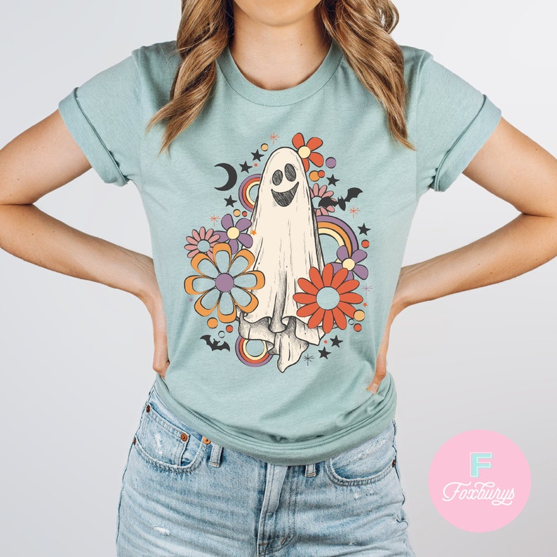 Peut inclure: Un t-shirt bleu clair avec un motif de fant&ocirc;me de style r&eacute;tro. Le fant&ocirc;me est blanc avec un visage souriant et est entour&eacute; de fleurs color&eacute;es, d'&eacute;toiles et de chauves-souris. Le design a une esth&eacute;tique vintage.