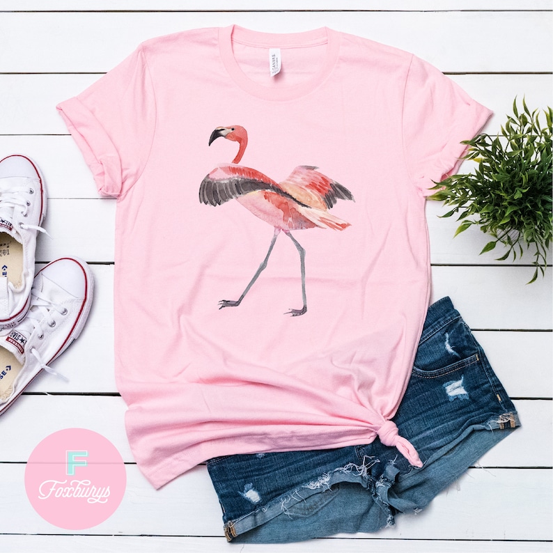 May include: Pink t-shirt with a watercolor flamingo graphic. The flamingo is standing with its wings spread out. The shirt is on a white wooden surface with a pair of white sneakers and a pair of denim shorts.