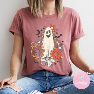 Peut inclure: Un t-shirt mauve avec un motif de fant&ocirc;me de style r&eacute;tro. Le fant&ocirc;me est entour&eacute; de fleurs color&eacute;es et d'&eacute;toiles. Le motif est imprim&eacute; sur le devant du t-shirt.