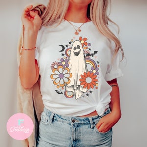 Peut inclure: T-shirt blanc avec un fant&ocirc;me souriant entour&eacute; de fleurs color&eacute;es et d'&eacute;toiles. Le fant&ocirc;me est blanc avec un contour noir et un sourire noir. Les fleurs sont orange, violette et rose avec des centres jaunes. Les &eacute;toiles sont blanches avec des contours noirs.