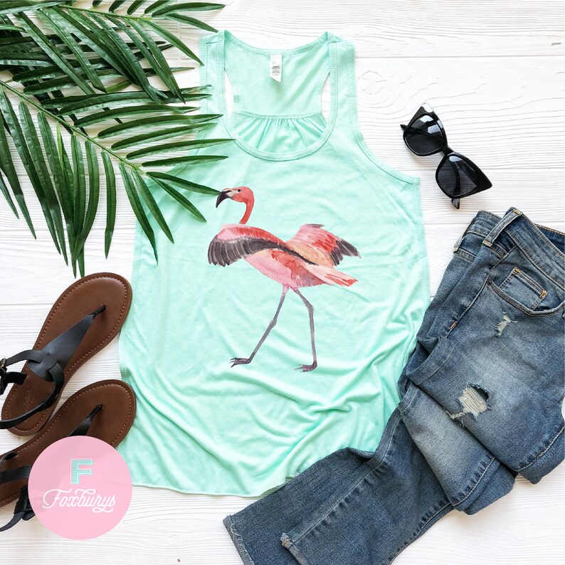 May include: A turquoise tank top with a pink flamingo graphic. The flamingo is standing on one leg with its wings spread out. The tank top is laying on a white wooden surface with a pair of sunglasses, a pair of brown sandals, and a pair of blue jeans.