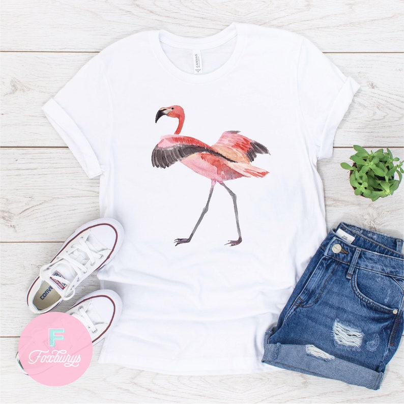 May include: White t-shirt with a pink flamingo illustration. The flamingo is standing on one leg with its wings spread out. The t-shirt is on a white wooden surface with a pair of white sneakers, blue denim shorts, and a small potted plant.