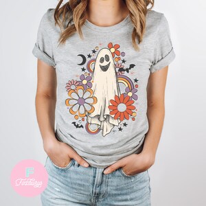Peut inclure: Un t-shirt gris avec un fant&ocirc;me blanc souriant entour&eacute; de fleurs color&eacute;es, d'&eacute;toiles et de chauves-souris. Le design a une ambiance r&eacute;tro des ann&eacute;es 70.